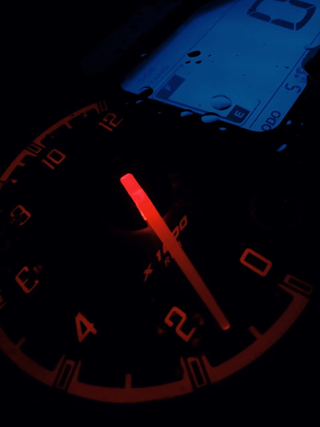 Red illuminated car tachometer with a glowing needle next to a blue digital ODO display behind water droplets.