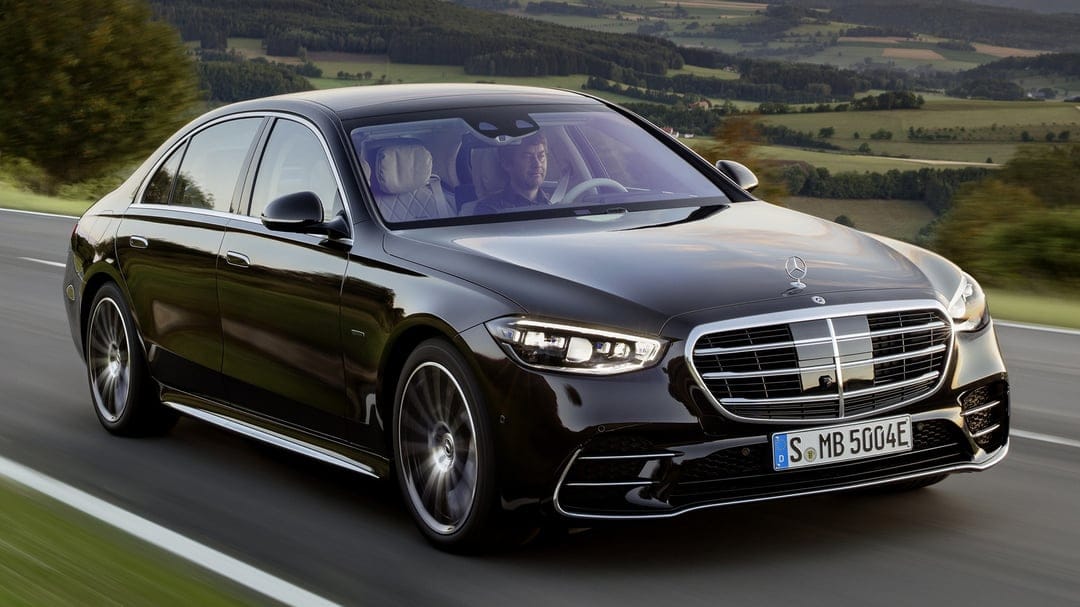 Black Mercedes-Benz S-Class luxury sedan driving on a scenic road with rolling hills in the background.