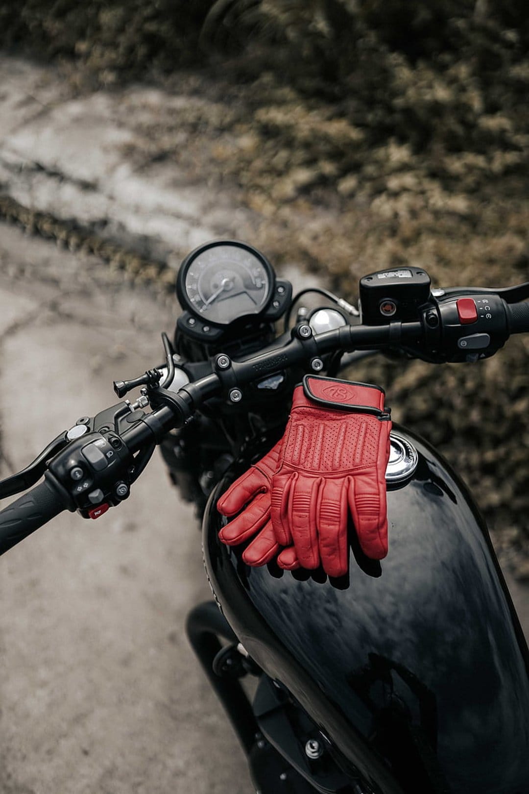 Red leather gloves on a black fuel tank with chrome handlebars and a round analog speedometer visible behind.
