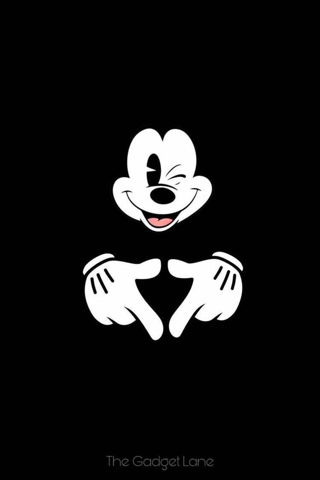 Mickey Mouse winks while forming a heart with his gloved hands against a solid black backdrop in a vertical layout.