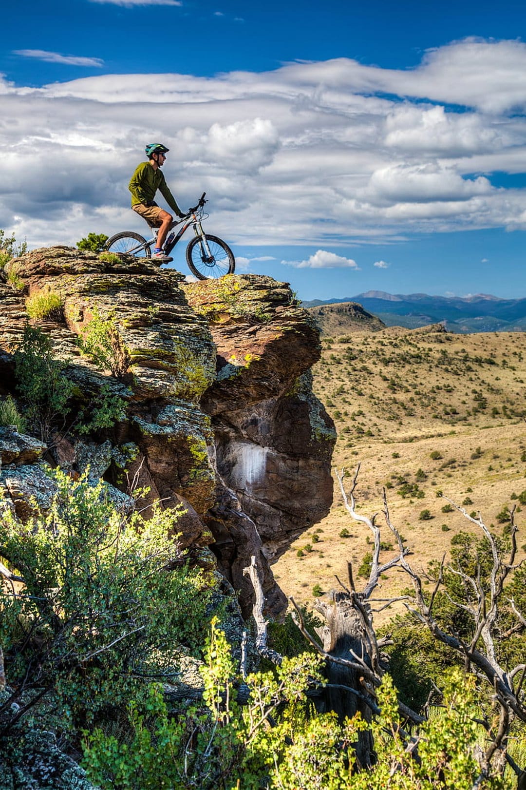 Mountain biker in a green shirt stands with his bike on a steep rocky cliff overlooking a vast desert valley.