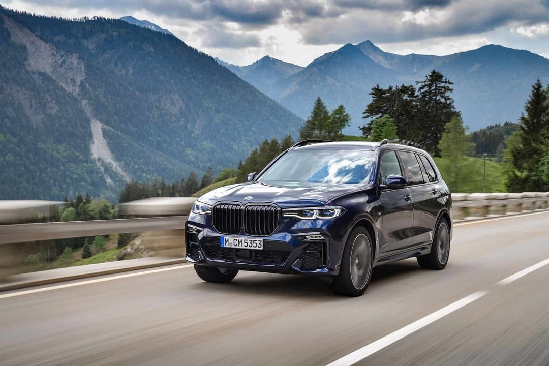 Dark blue luxury SUV driving on a mountain road with a backdrop of hazy peaks and green forests under a cloudy sky.