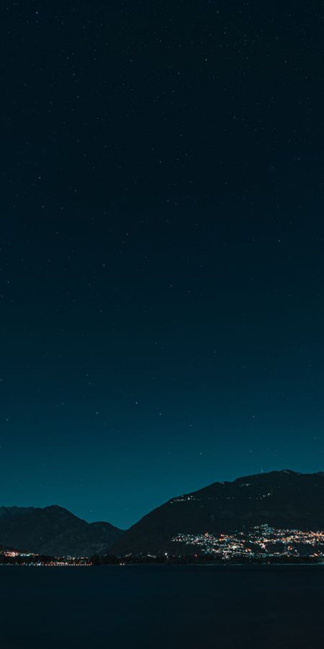 Glowing village lights reflect in a dark lake beneath a vast starry sky and distant mountain silhouettes.