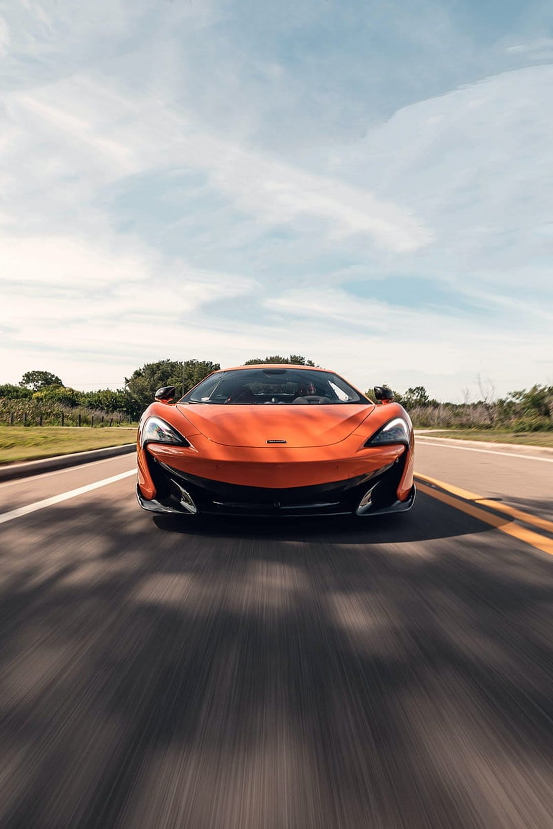 Orange McLaren supercar speeds along a tree-lined road under a bright sky with visible motion blur effects.