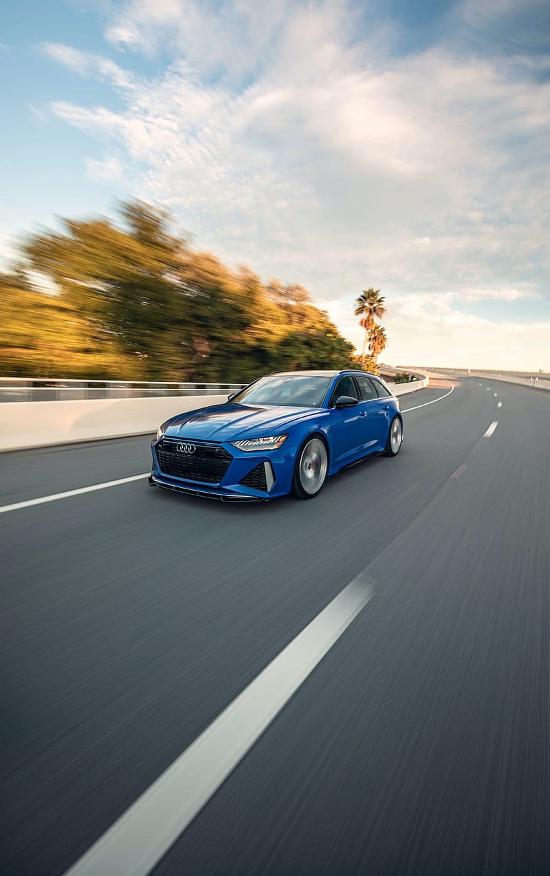 Blue Audi RS6 Avant sports wagon driving on a paved road lined with green trees and palms under a bright sky.