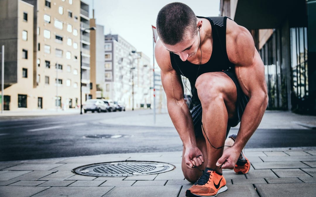 Muscular man in a black tank top ties orange running shoes on a city sidewalk with buildings in the background.