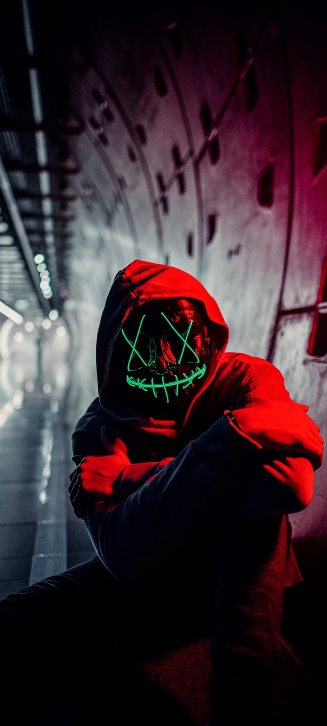 Man in a red hoodie wearing a glowing green LED mask sits in a dimly lit tunnel with urban red lighting.
