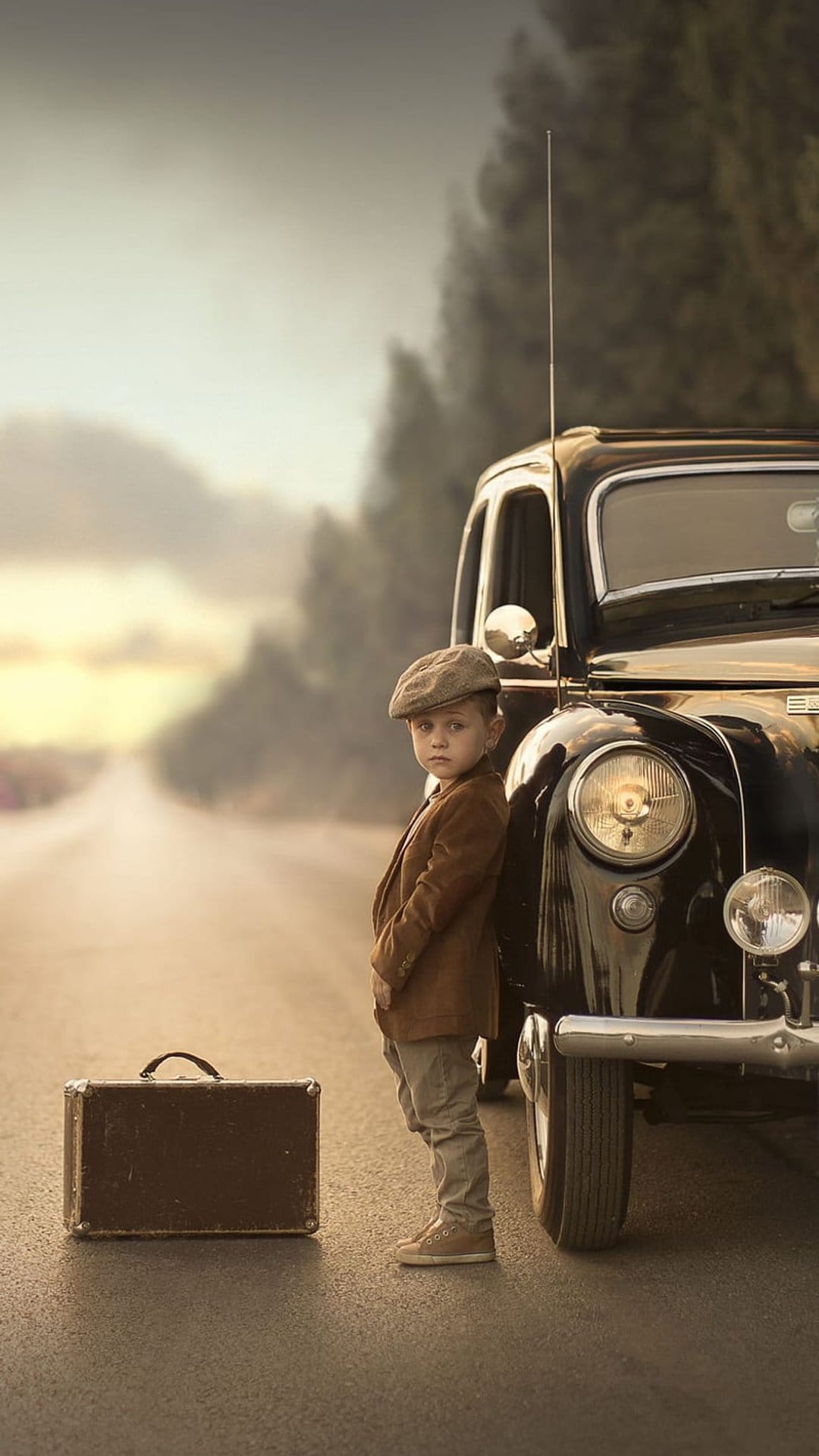 Young boy in a flat cap stands by a black vintage car and suitcase on a hazy, golden country road.
