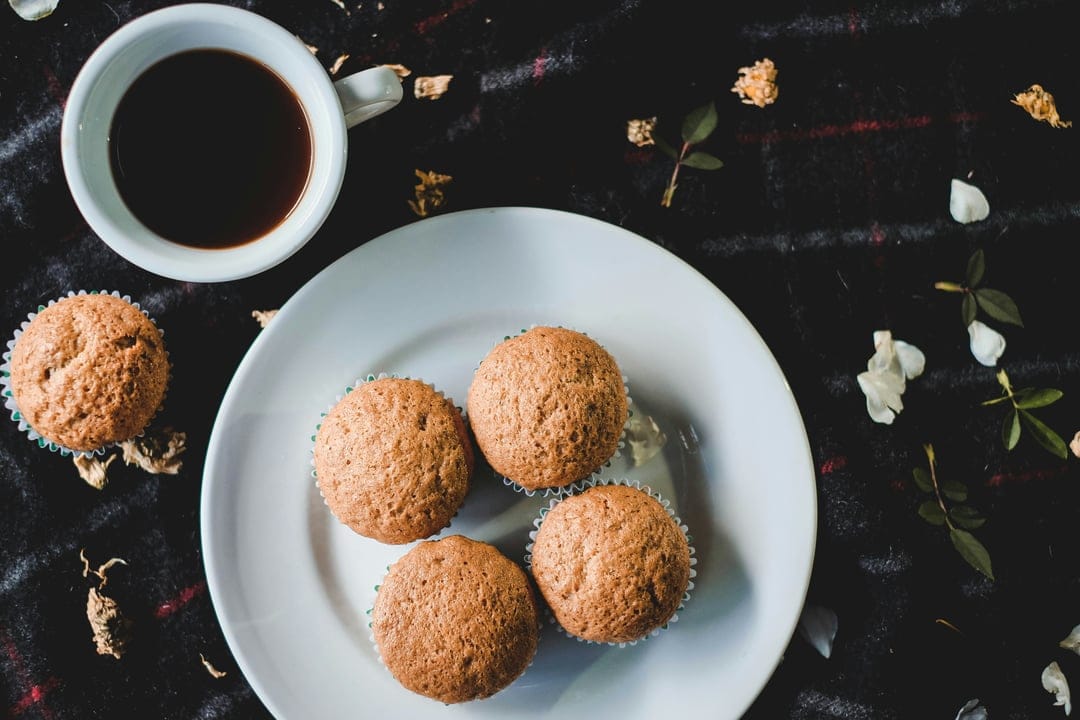 Four muffins on a white plate next to a cup of coffee and dried flowers over a dark textured surface.