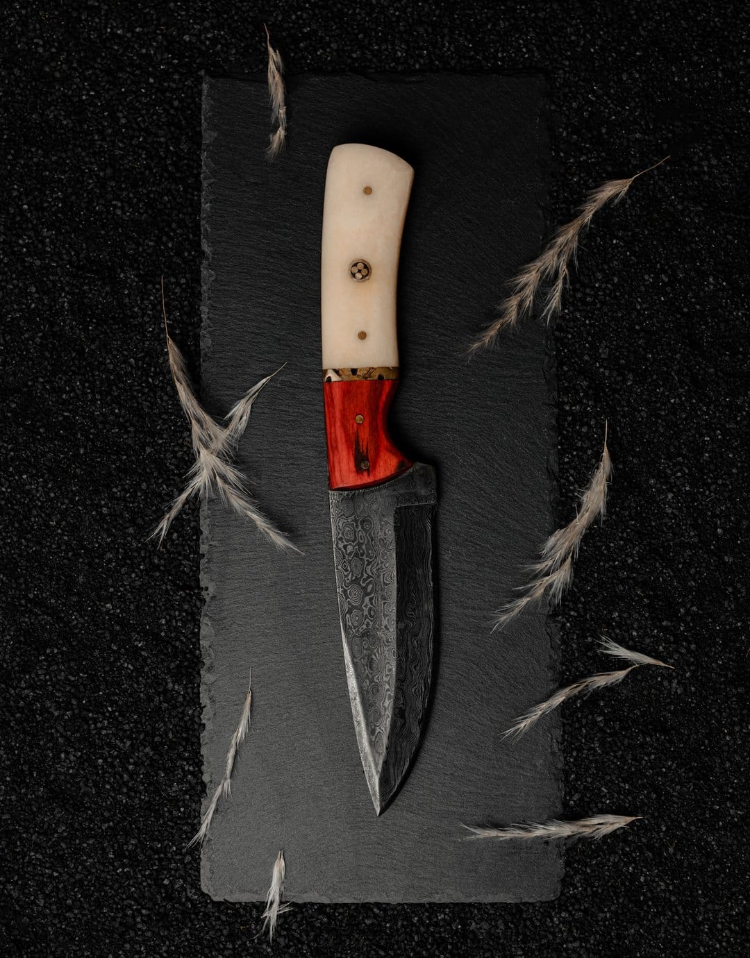 Damascus steel knife with a carved bone handle and red accents lies on dark slate near wisps of dried grass.