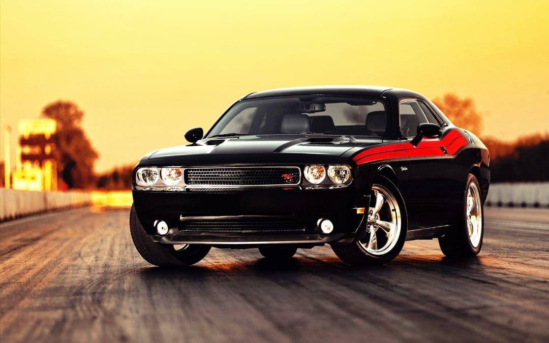 Black Dodge Challenger R/T with red racing stripes on a sunset drag strip with warm golden hour lighting.