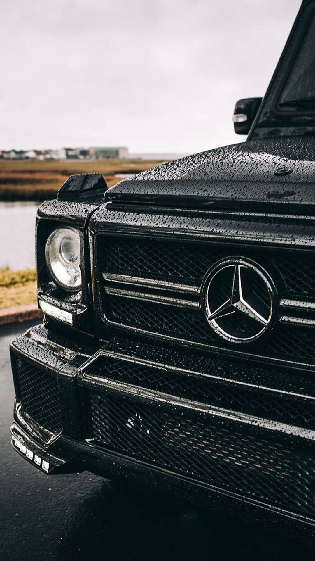 Black Mercedes G-Wagon front grille with raindrops against a misty lake background on a rainy day.