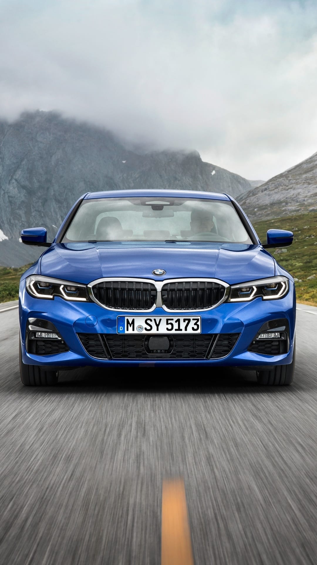 Front view of a blue BMW sedan driving on a mountain road with misty peaks and a cloudy sky background.
