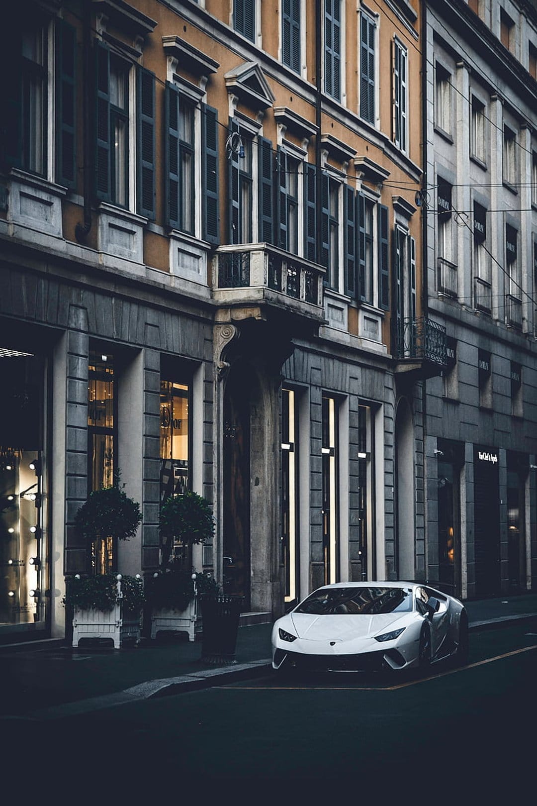 White Lamborghini parked on a dim European street in front of a classic stone building with ornate windows.