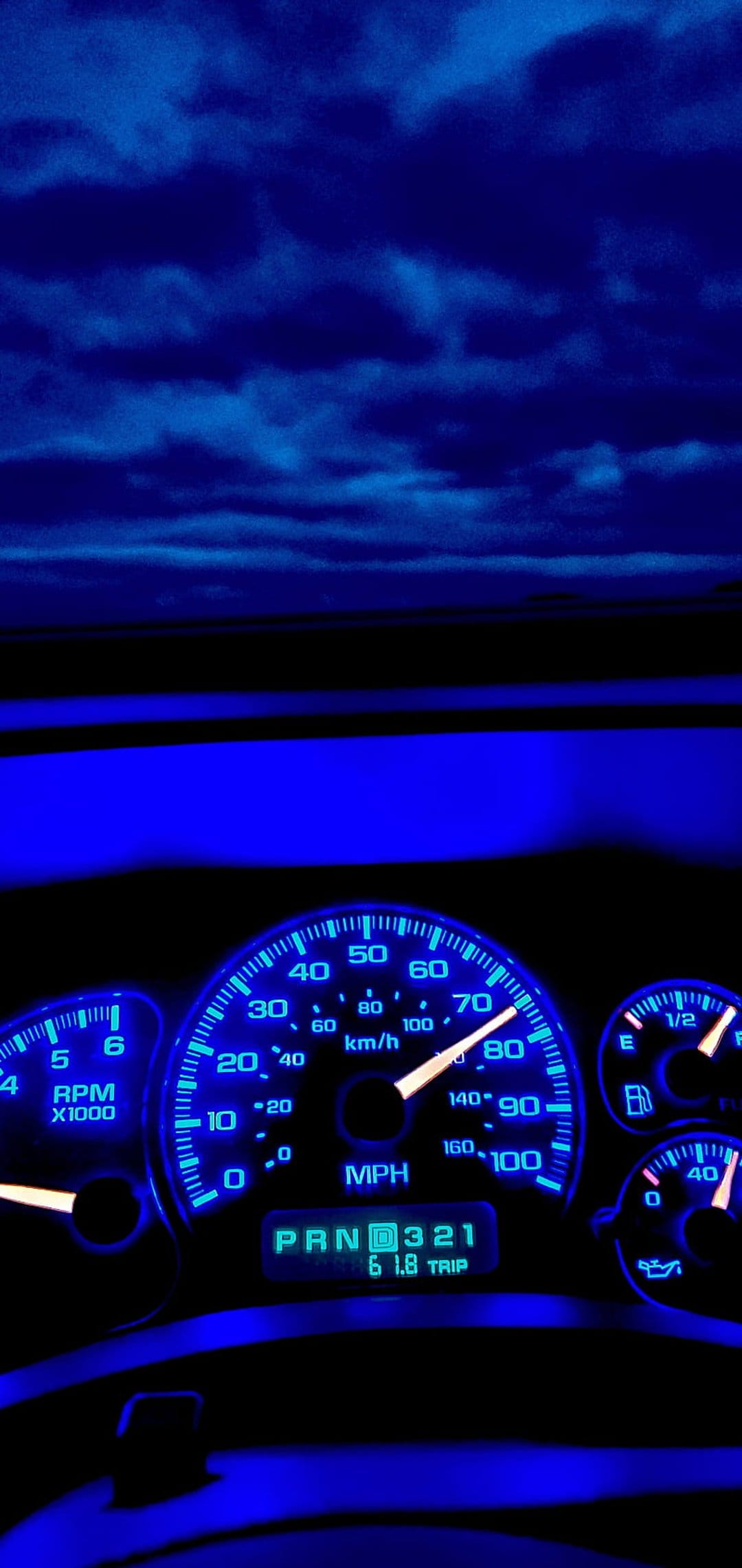 Illuminated blue speedometer and tachometer gauges on a car dashboard against a twilight sky background.