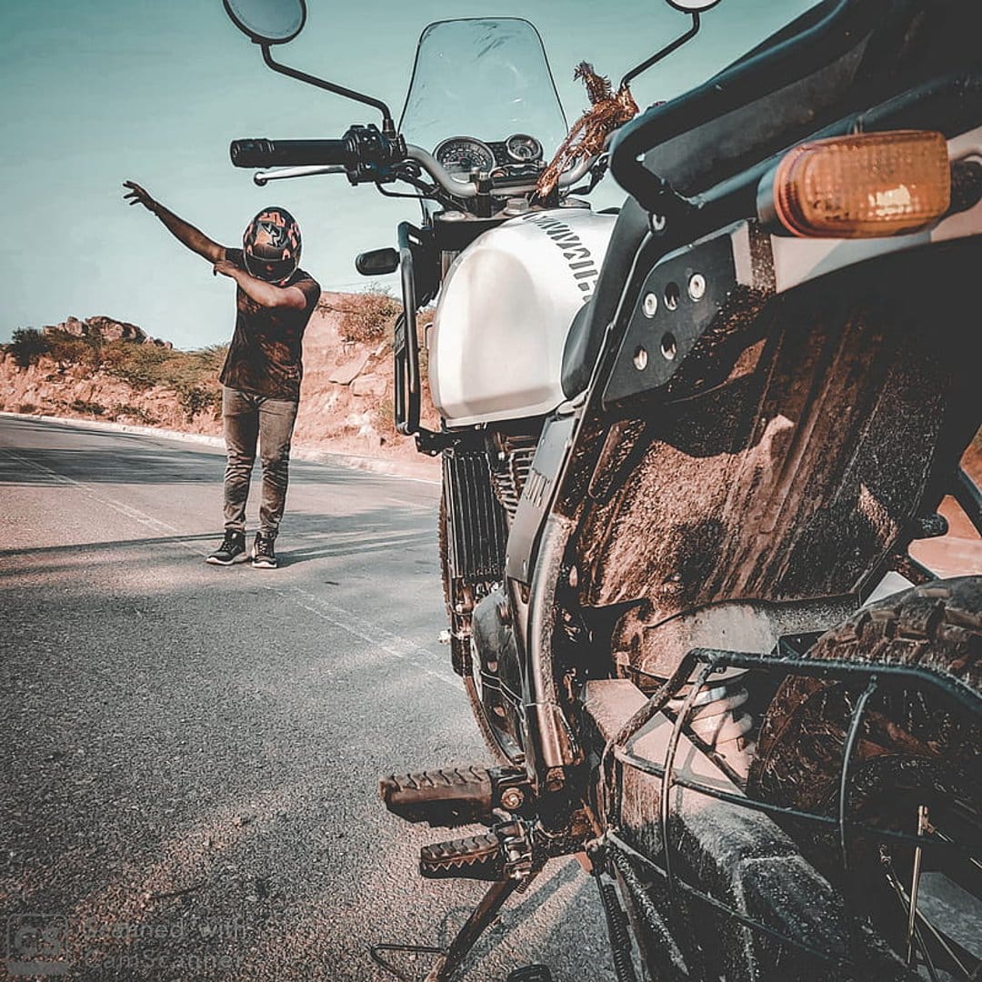 Man in a helmet gestures next to a white motorcycle on a dusty desert road with rocky hills under a blue sky.