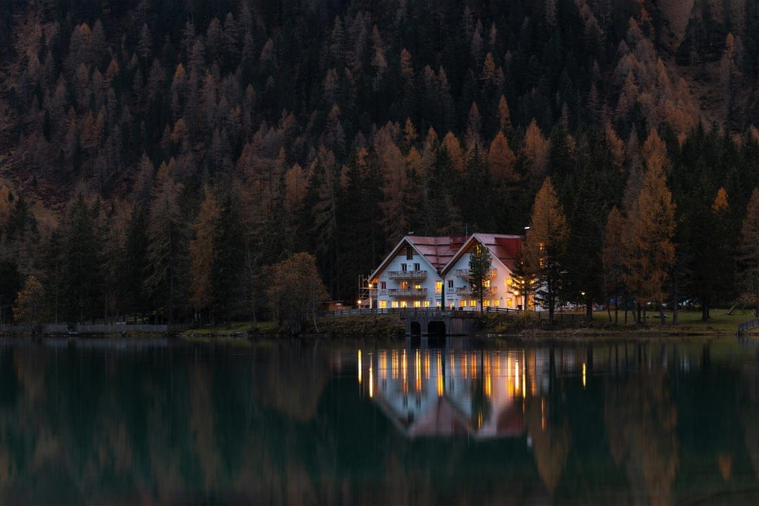 Wooden cabin with glowing windows reflects in a still lake surrounded by dense pines under a dusky evening sky.