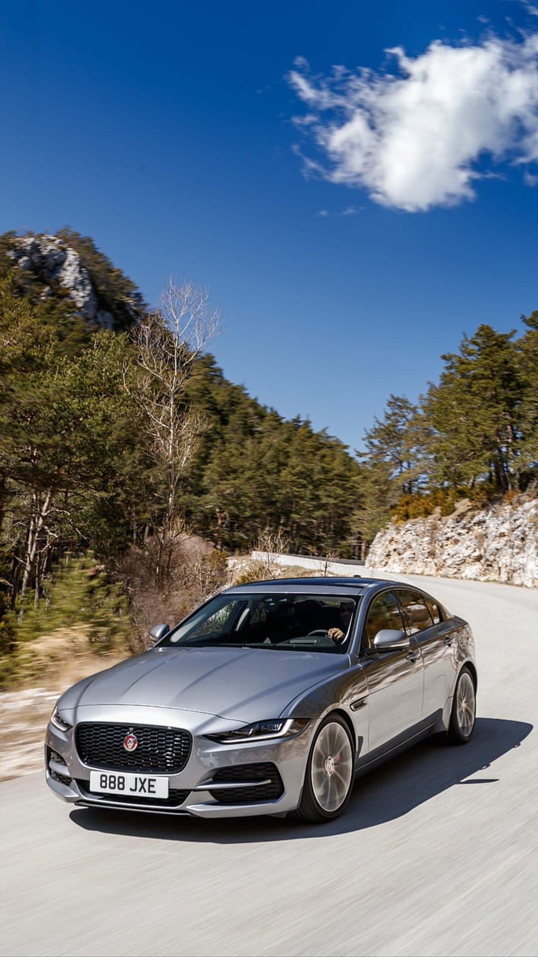 Silver Jaguar XE sedan drives along a winding mountain pass with lush green forests under a bright blue sky.