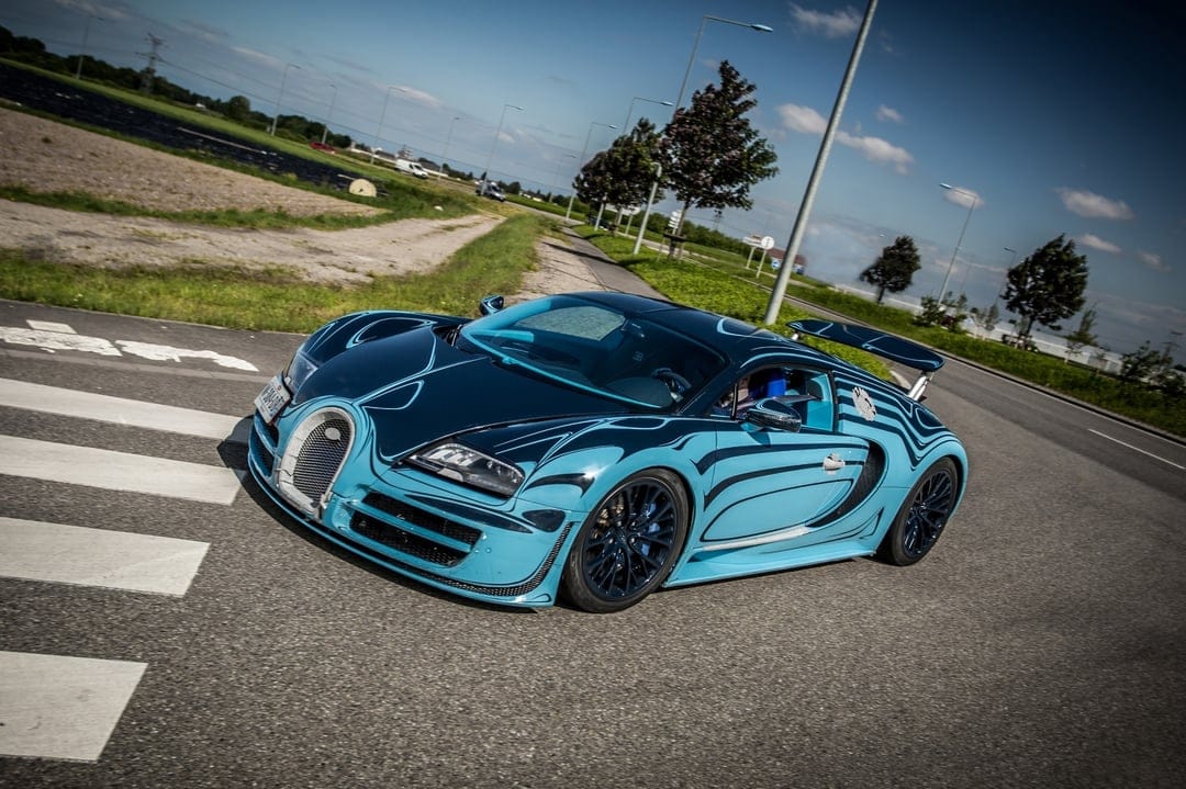 Blue Bugatti Veyron hypercar with flowing paint patterns driving on a sunny road under a clear sky.