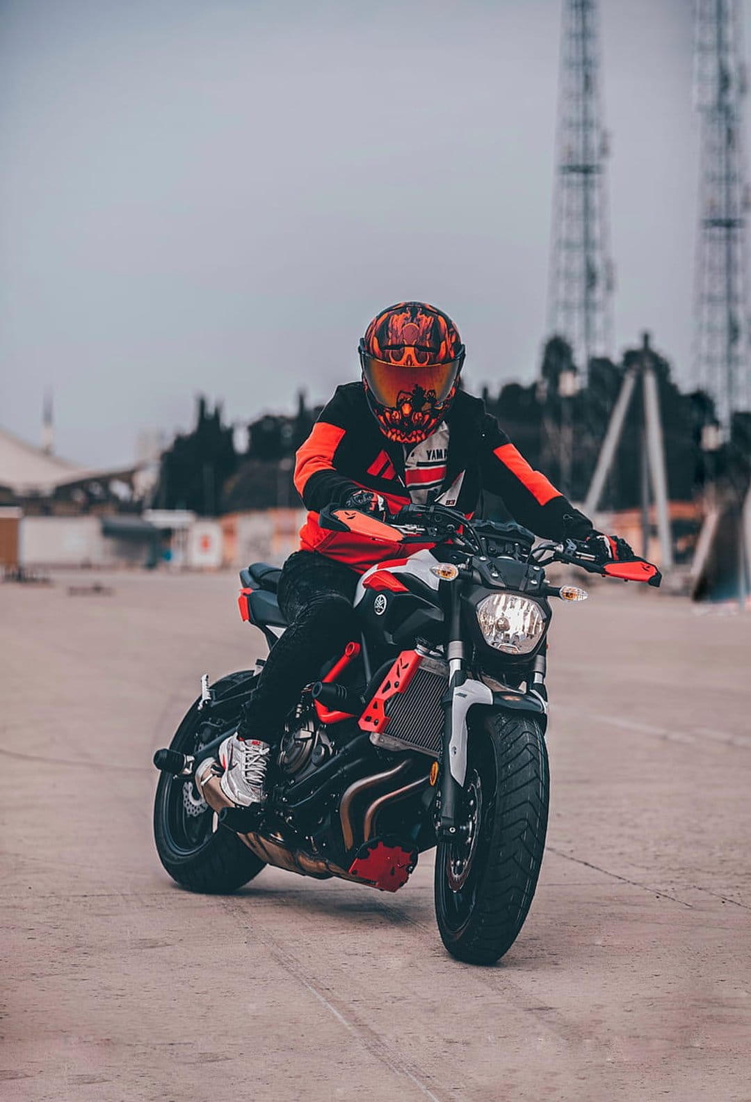 Motorcyclist in orange and black riding gear sits on a Yamaha motorcycle against an industrial urban backdrop.
