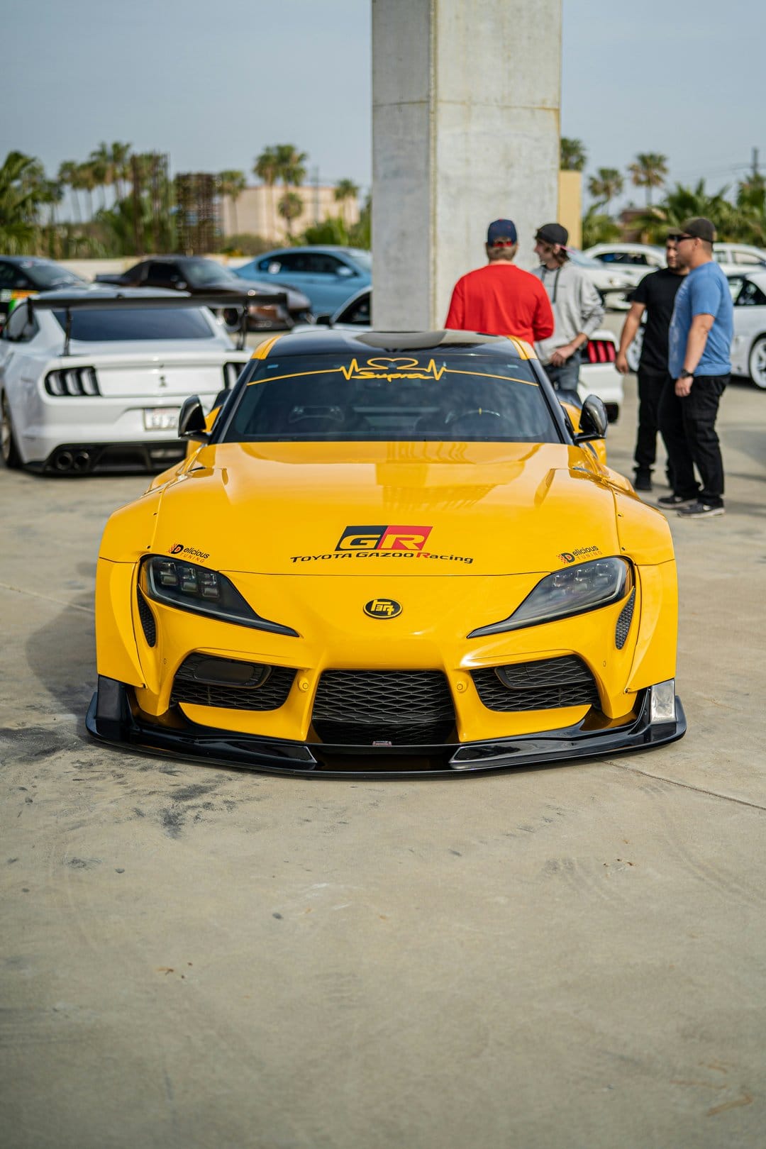 Yellow Toyota Supra GR with widebody kit and Gazoo Racing decals parked at a sunny car meet near palm trees.