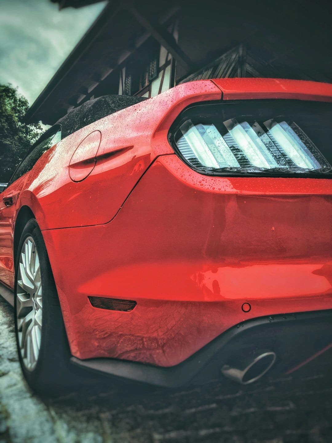 Red Ford Mustang rear view covered in raindrops with glowing taillights against a blurred urban background.