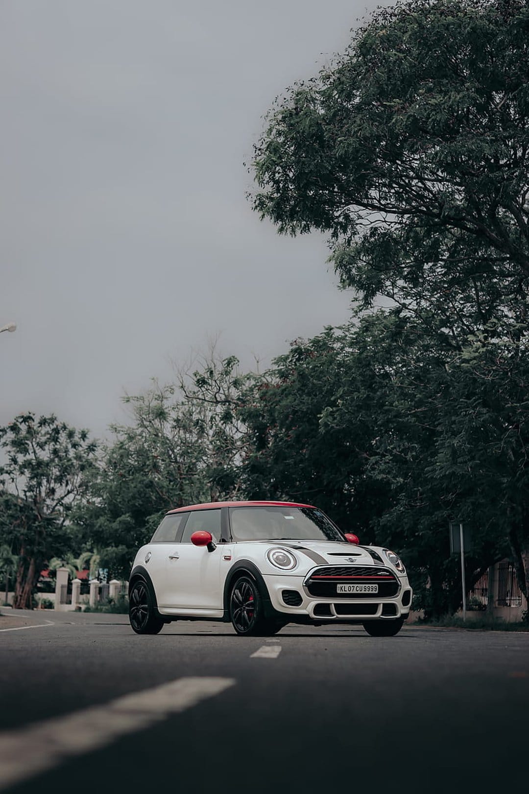 White Mini Cooper with a red roof and racing stripes parked on a paved road surrounded by green trees.