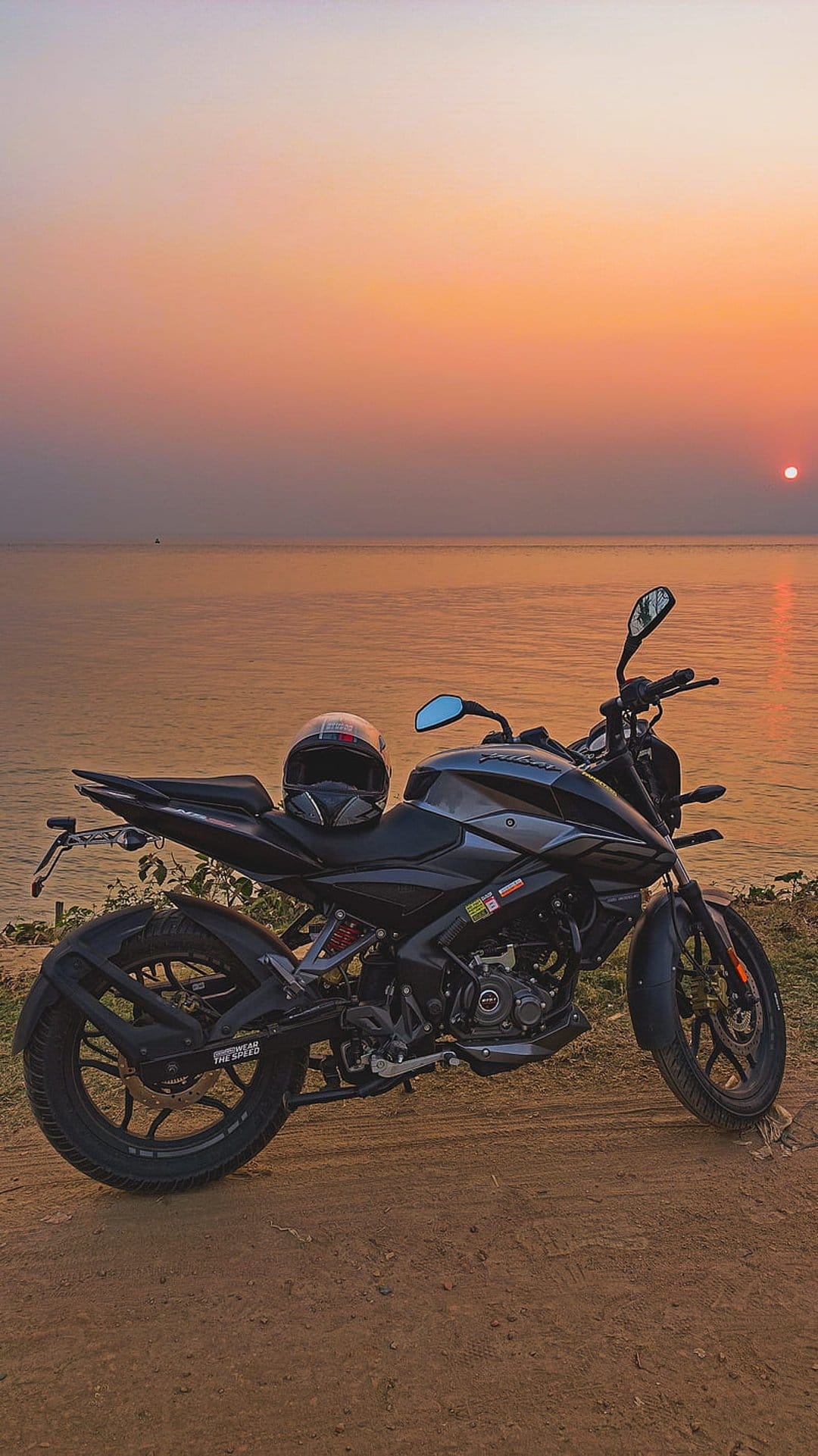 Black motorcycle with a helmet on the seat parked on a coastal cliff path during a vibrant orange sunset sky.