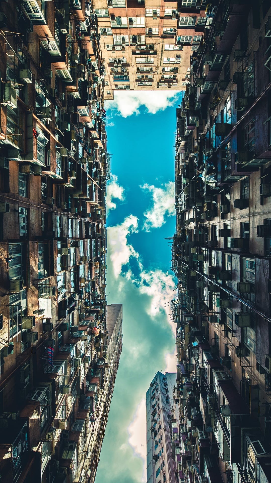 High-rise apartment buildings with air conditioning units frame a narrow strip of blue sky and wispy white clouds.
