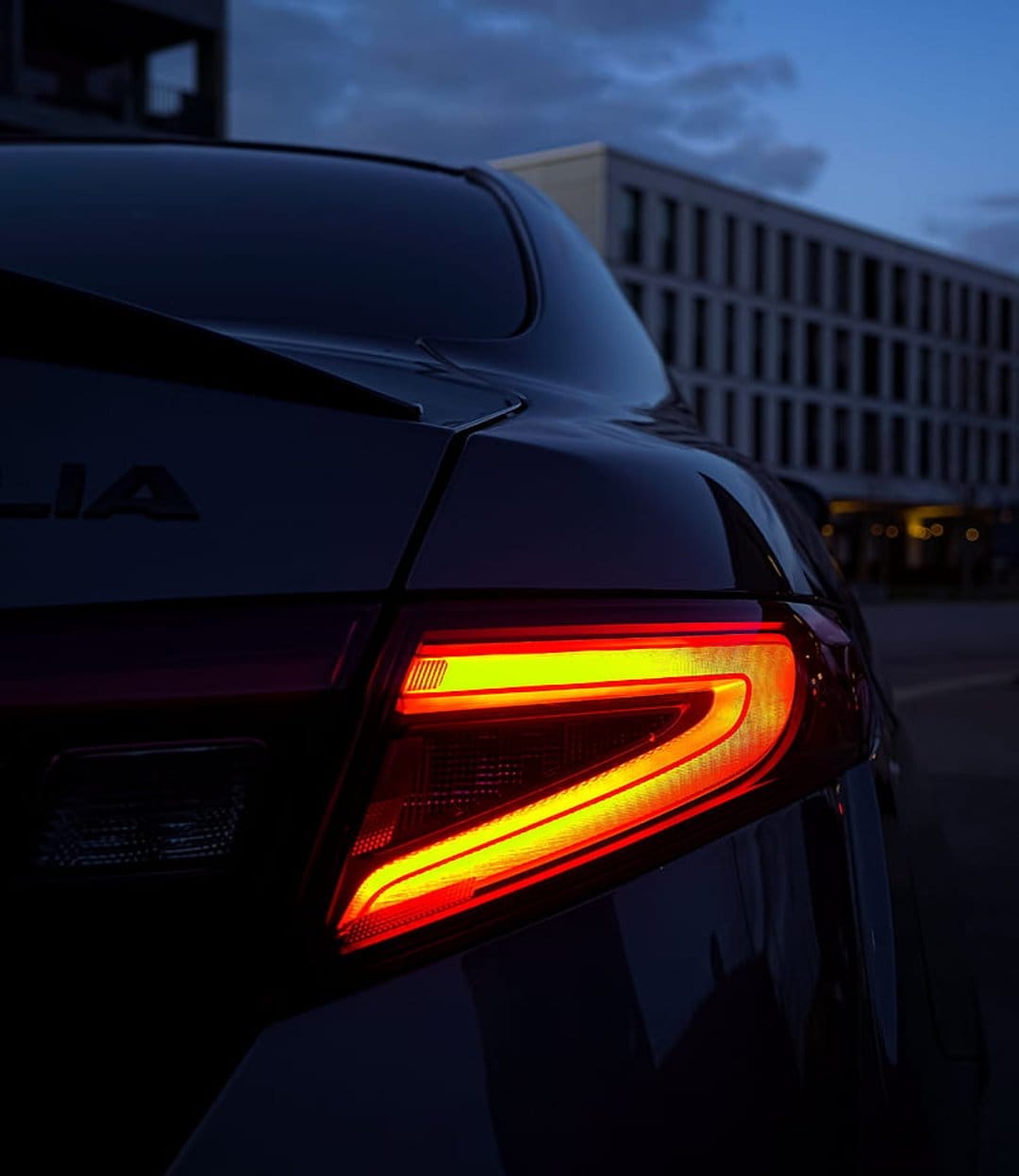 Red LED car tail light glows intensely at dusk against a minimalist urban building with lit windows.