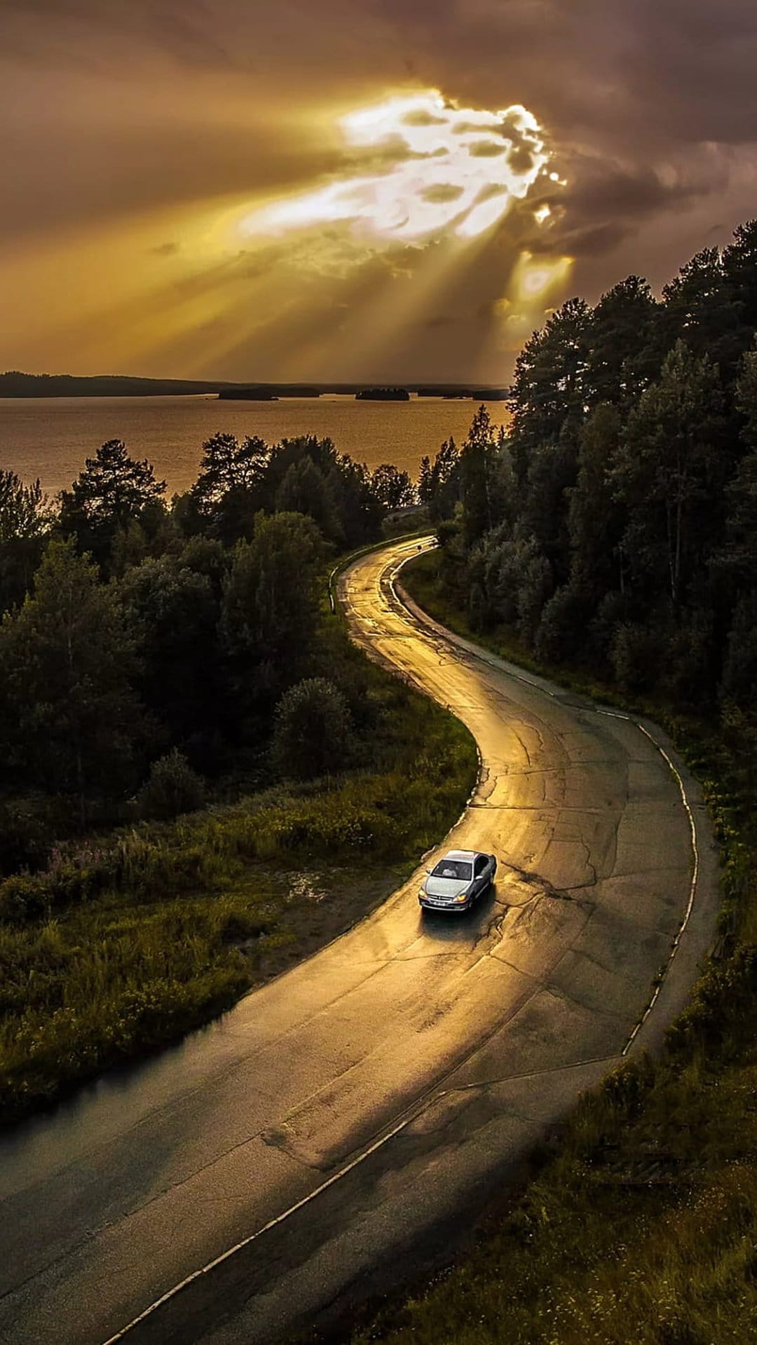 Silver car drives on a wet winding road through a forest as sunbeams pierce through dark clouds over a lake.