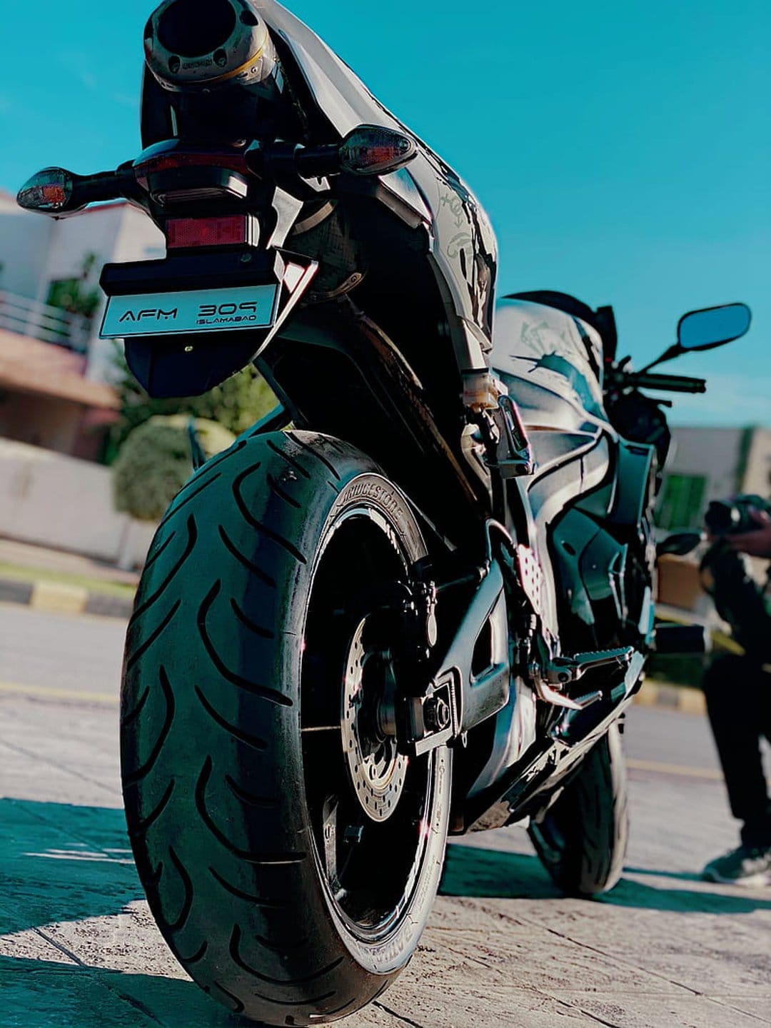 Sportbike rear view showing a wide tire, glowing taillight, and license plate AFM 309 against a bright blue sky.