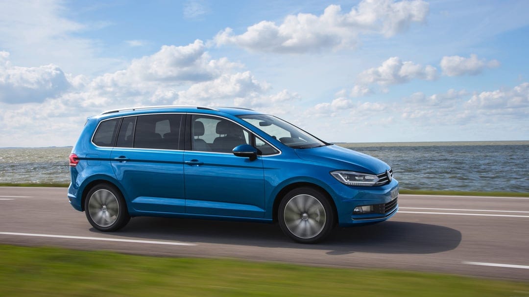 Blue Volkswagen Touran driving on a coastal road next to the ocean under a bright cloudy sky during daytime.