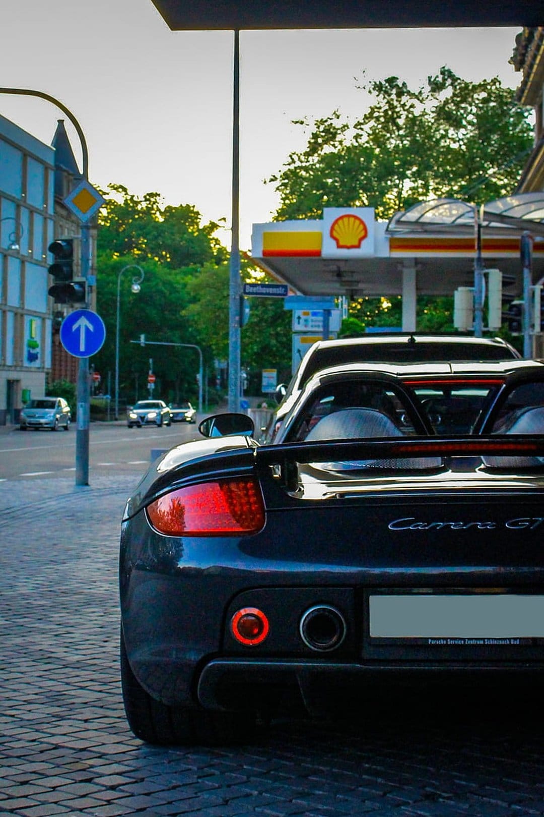 Porsche Carrera GT rear showing exhaust pipes and taillights on a cobblestone street by a Shell station.