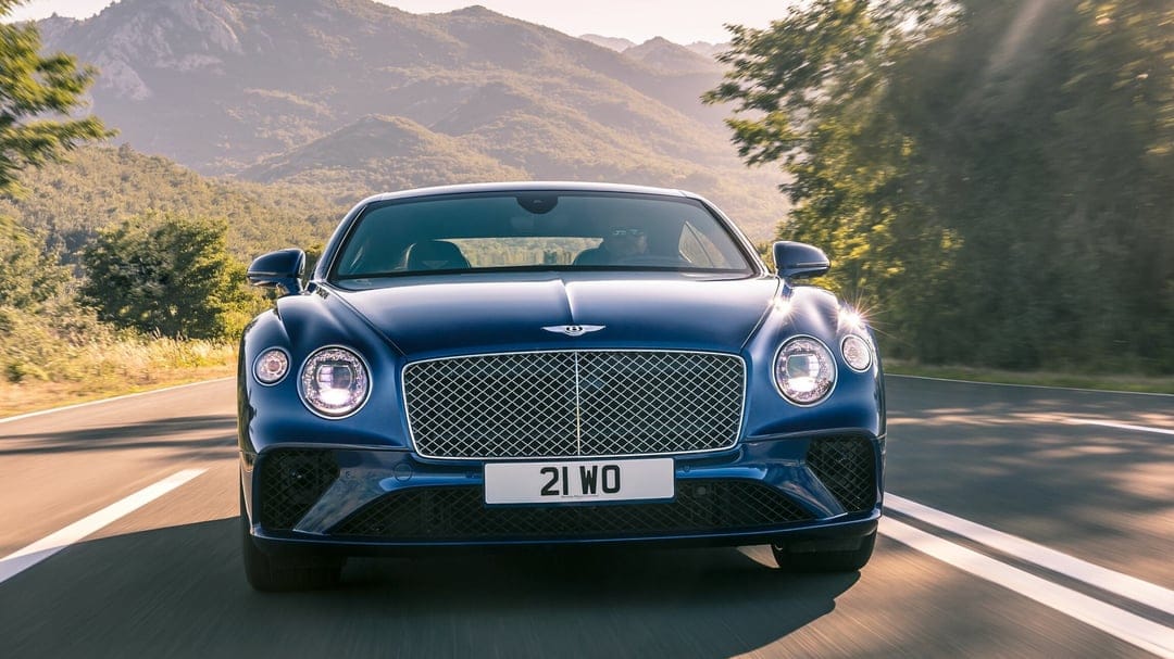 Blue Bentley Continental GT driving on a mountain road with warm sunlight and hazy peaks in the background.