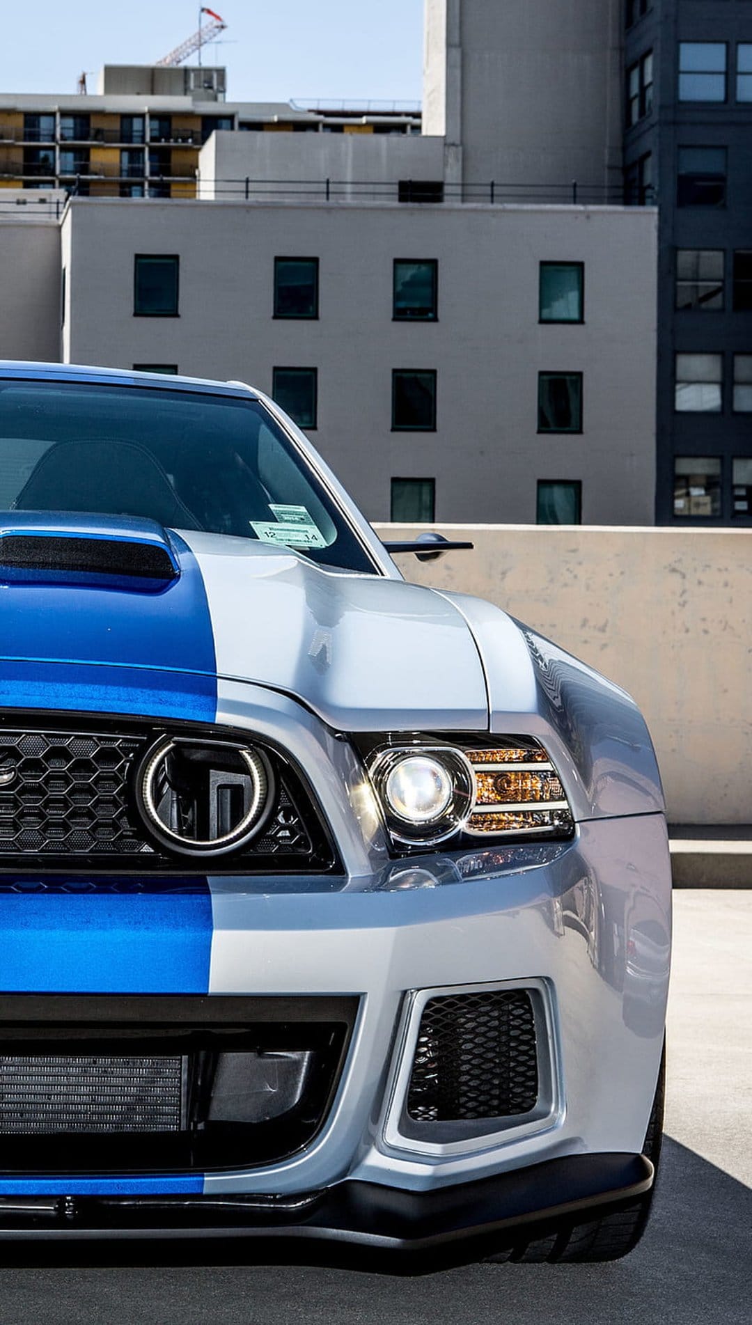 Ford Mustang front grille and LED headlight with blue racing stripes against a modern city building backdrop.
