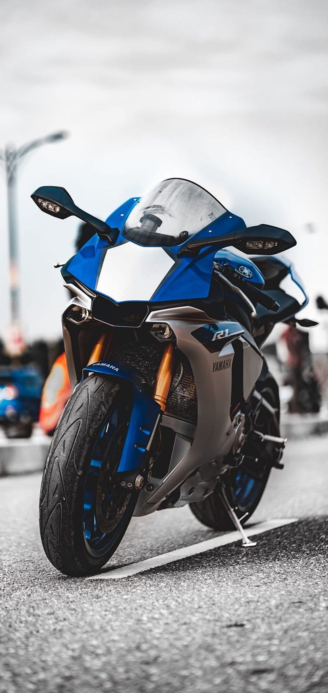 Blue Yamaha R1 motorcycle with golden forks parked on an asphalt street with a blurred urban city background.