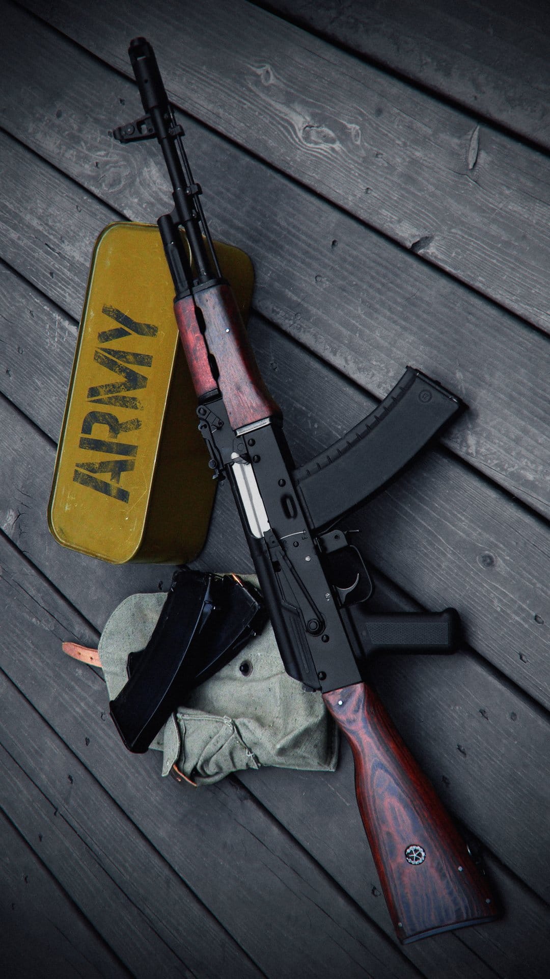 AK-47 rifle with a wooden stock lies next to a yellow ammunition crate and camouflage cap on a dark table.