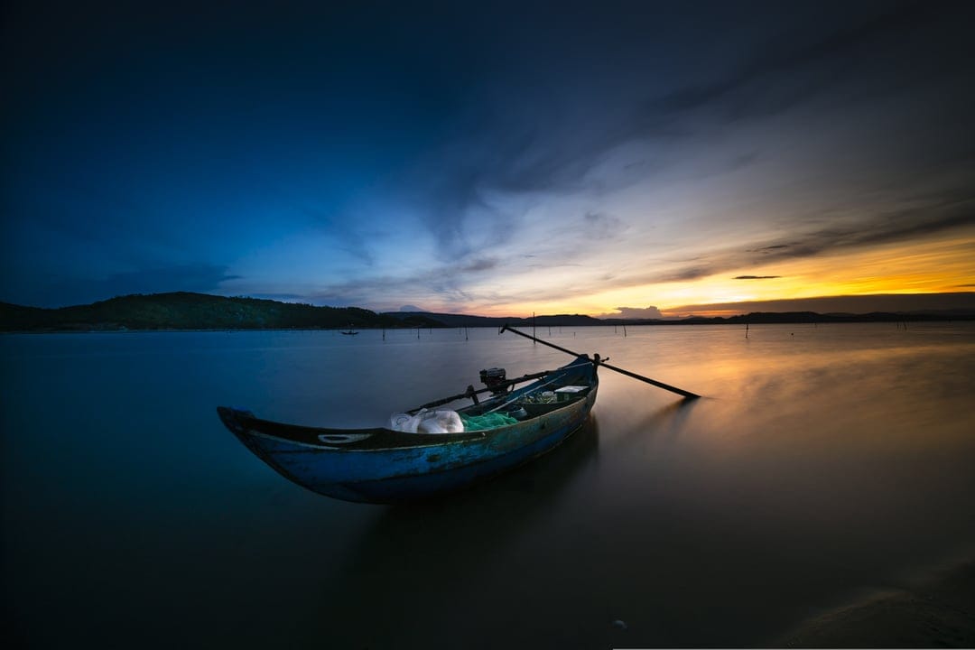 Blue fishing boat floating on smooth water during a vibrant orange and purple sunset with a soft hazy horizon.