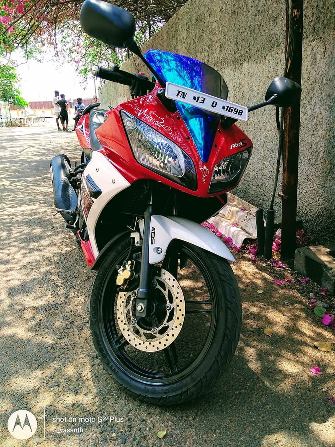 Red Yamaha R15 motorcycle with a blue wind deflector parked on gravel with pink petals under bright sunlight.