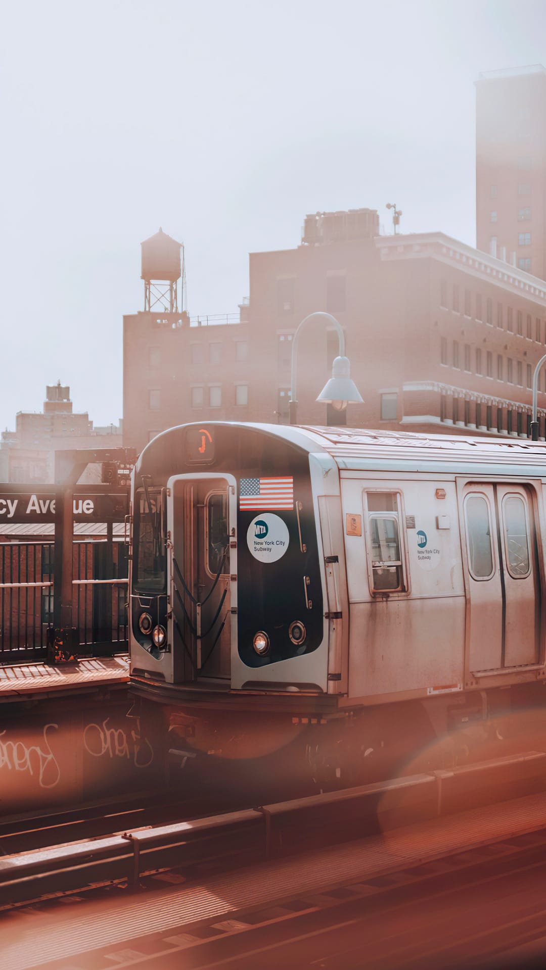 Subway train with an American flag emblem sits at a misty station platform under a hazy city skyline at dawn.