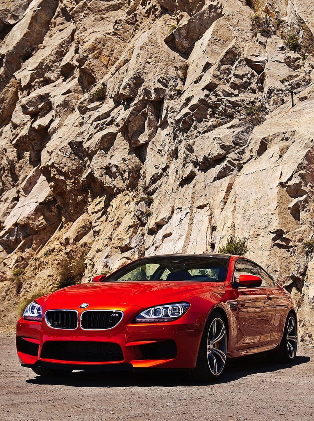 Red BMW M6 Coupe parked on a dusty dirt road in front of a massive textured mountain rock face at a low angle.