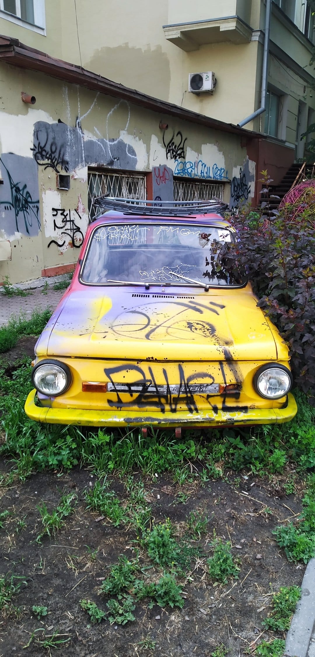 Yellow car with colorful graffiti sits in tall green grass before an abandoned urban building background.