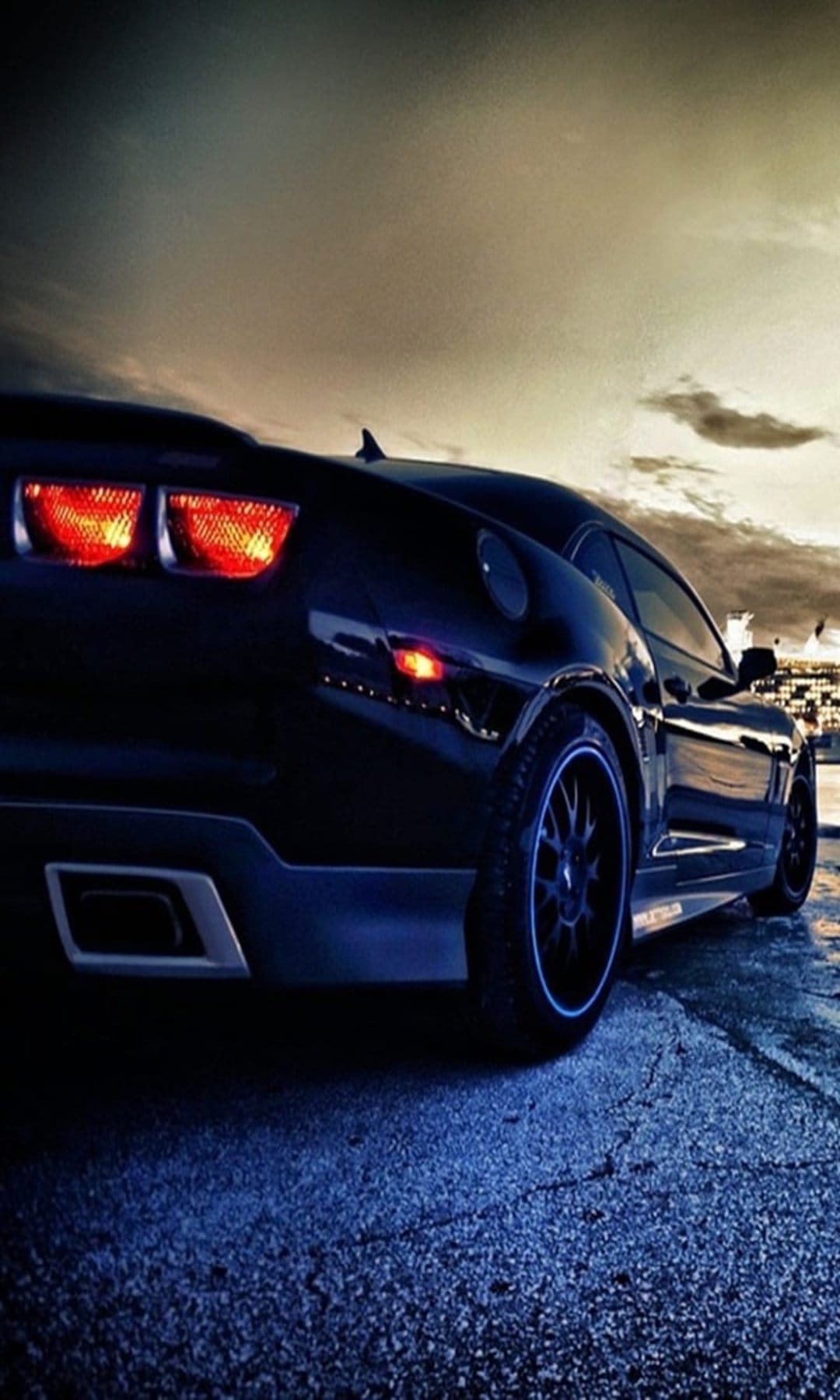 Black Chevrolet Camaro rear with glowing red tail lights under a moody dusk sky on textured asphalt ground.