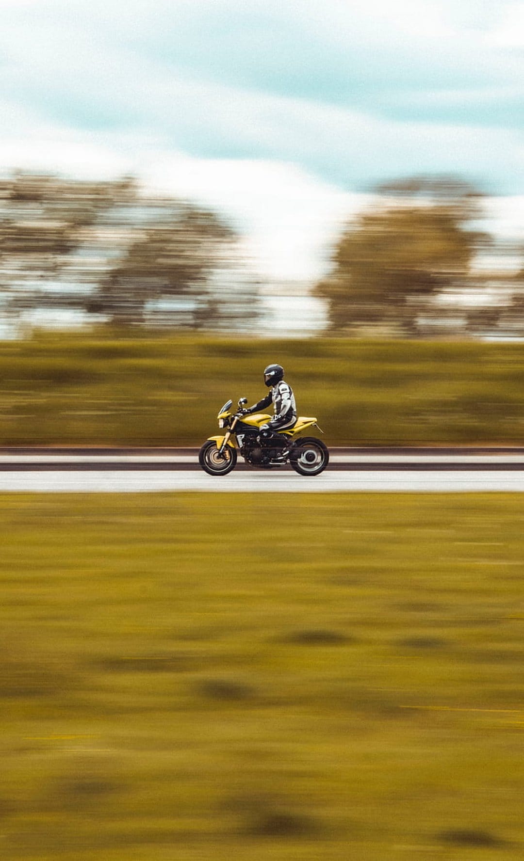 Motorcyclist wearing yellow gear riding fast on a road with motion blurred green trees and a soft blue sky.