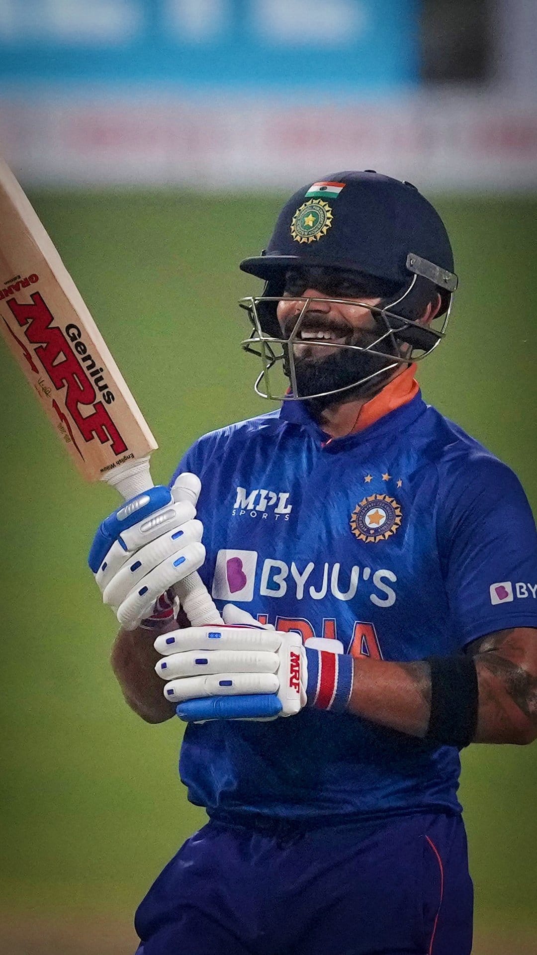 Virat Kohli smiles while holding an MRF cricket bat in a blue jersey and helmet during an outdoor match.