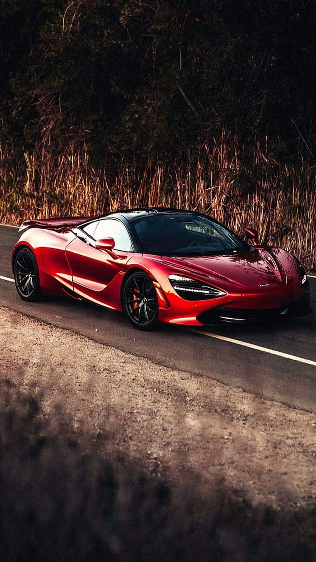 Red McLaren 720S supercar driving on a scenic road surrounded by dry grass and lush trees at a low angle.