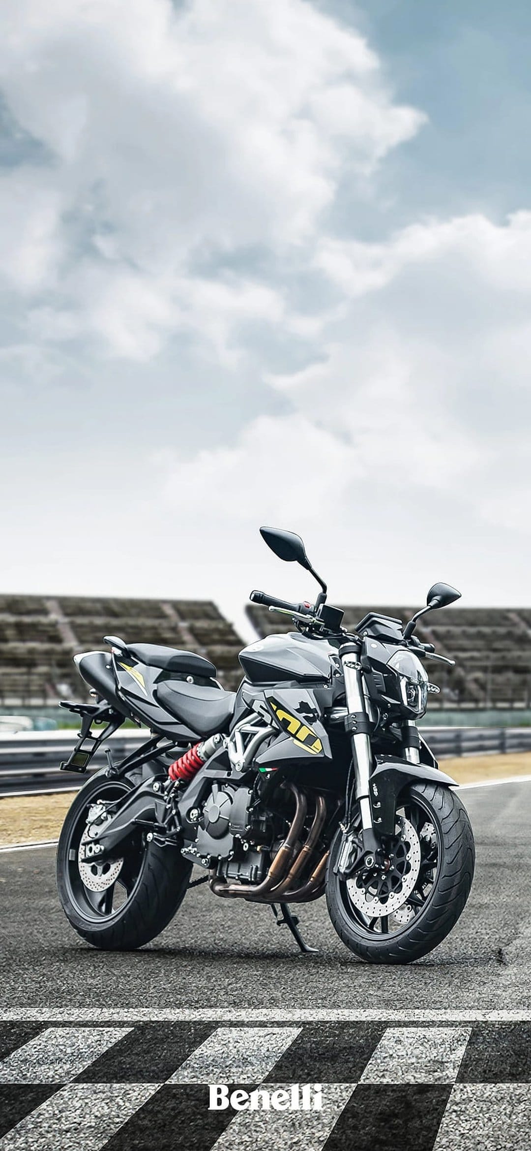 Black Benelli motorcycle on a racetrack with a red shock absorber, blurred stadium, and cloudy sky overhead.
