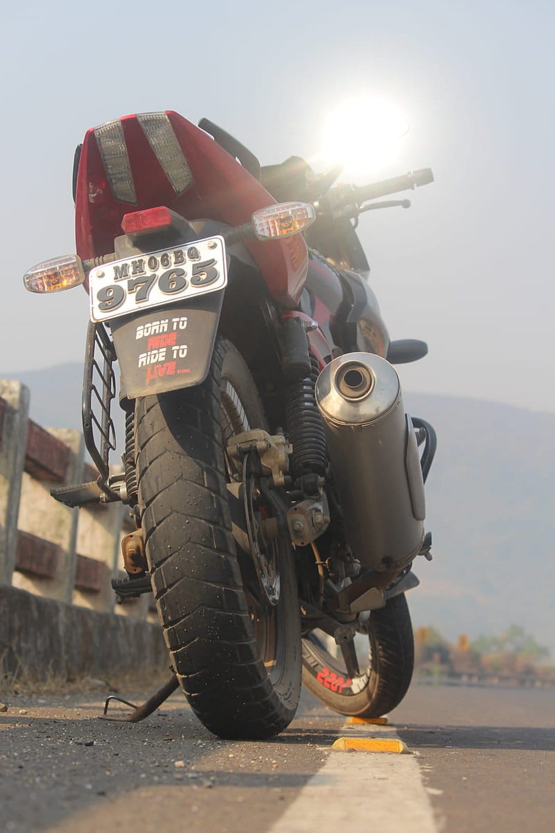 Motorcycle rear tire and exhaust pipe on a mountain road under a bright sun lens flare with a quote sticker.
