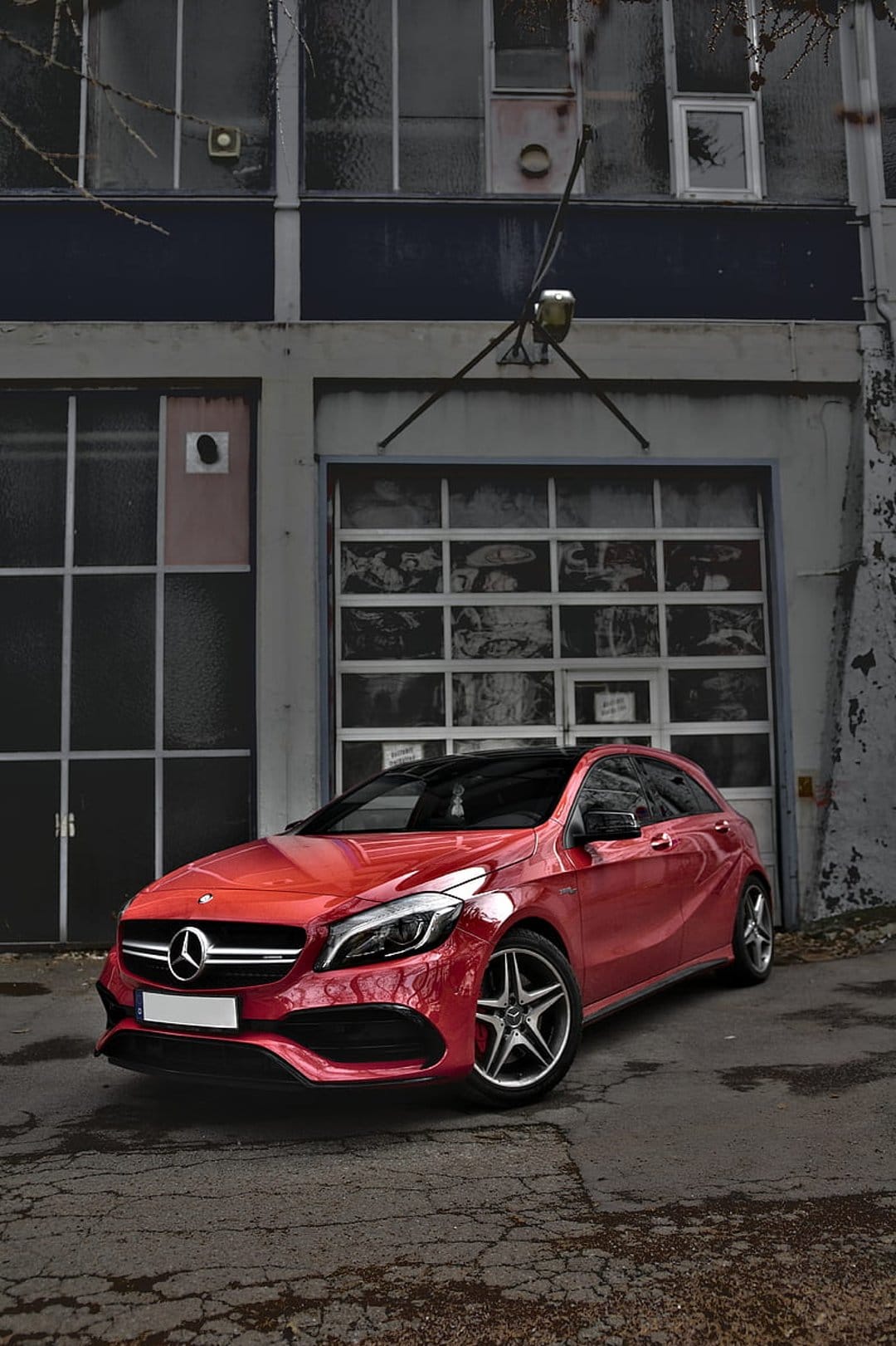 Crimson Mercedes-Benz A-Class hatchback parked before a large industrial garage door at an urban site.